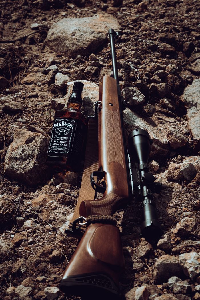 A bottle of Jack Daniels whisky next to a rifle laid on rocky terrain.