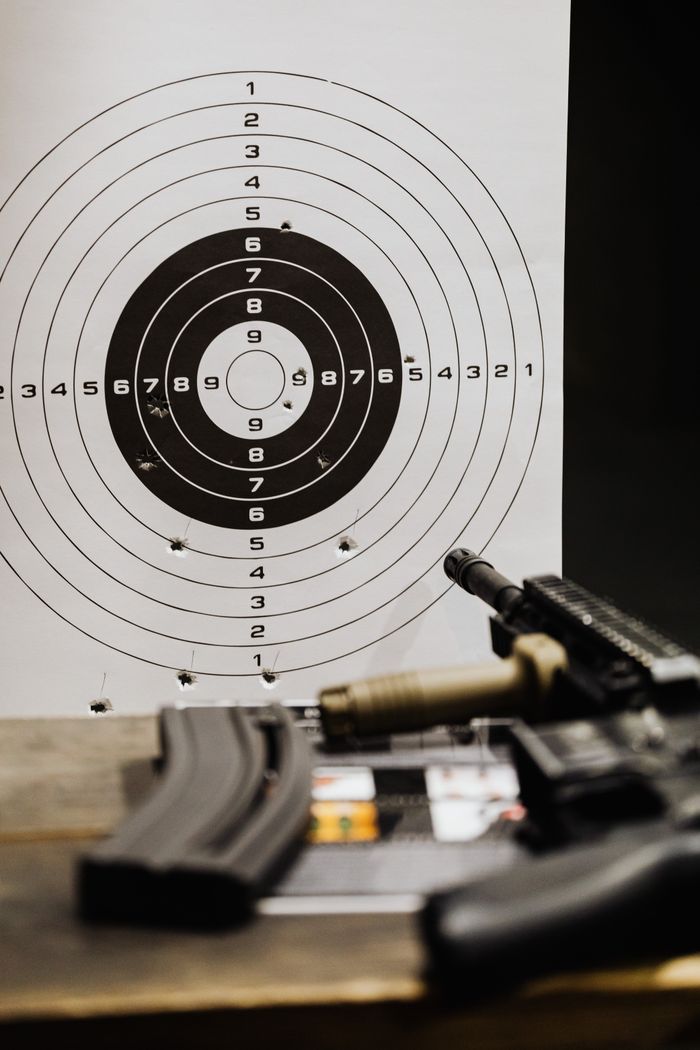 contact-img Close-up of a rifle resting on a wooden surface with a target paper in the background.